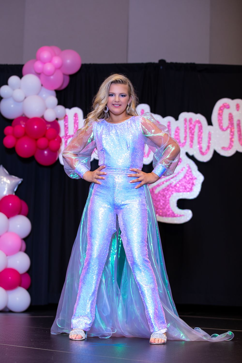 Model wearing iridescent jumpsuit with sheer overlay poses on stage with pink balloon decorations.