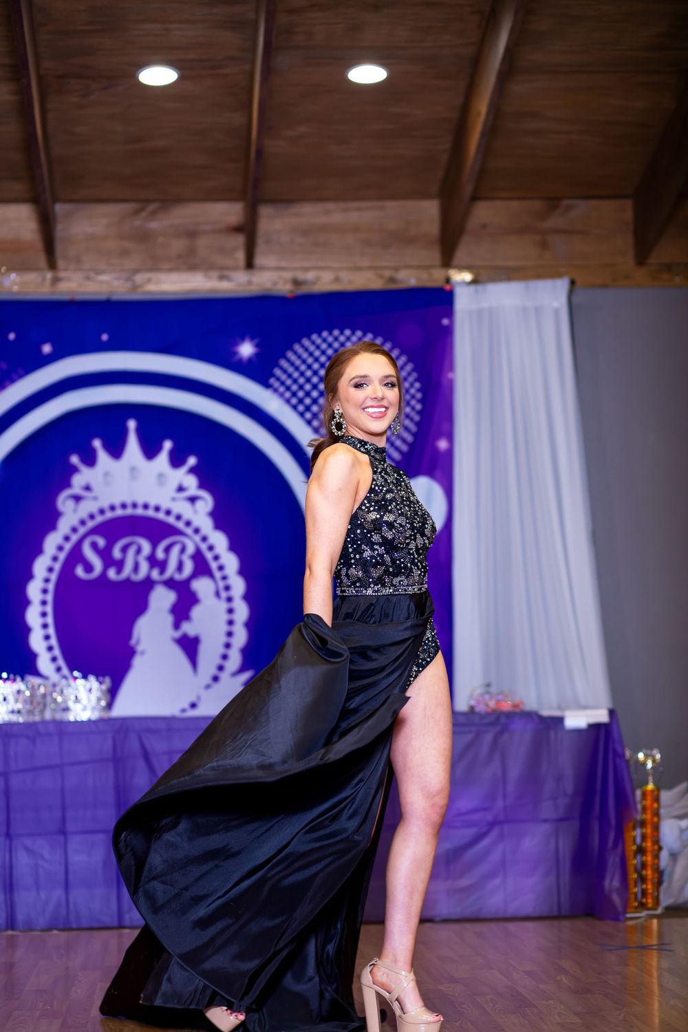 Elegant model poses in a black gown with beaded top and flowing skirt on stage with blue and purple backdrop.