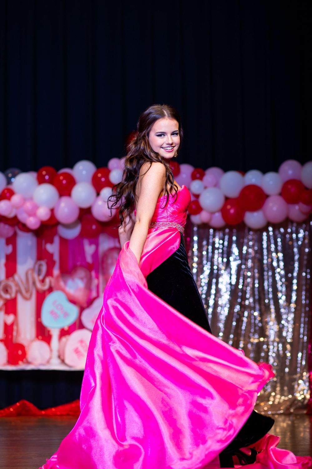 Performer twirls in flowing hot pink gown on stage with silver and balloon decorations.