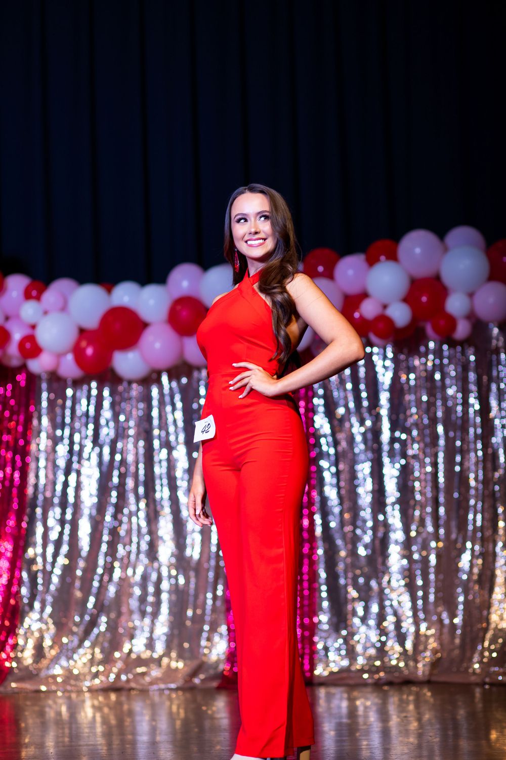 Model strikes confident pose in red formal jumpsuit against sparkly backdrop with balloons.