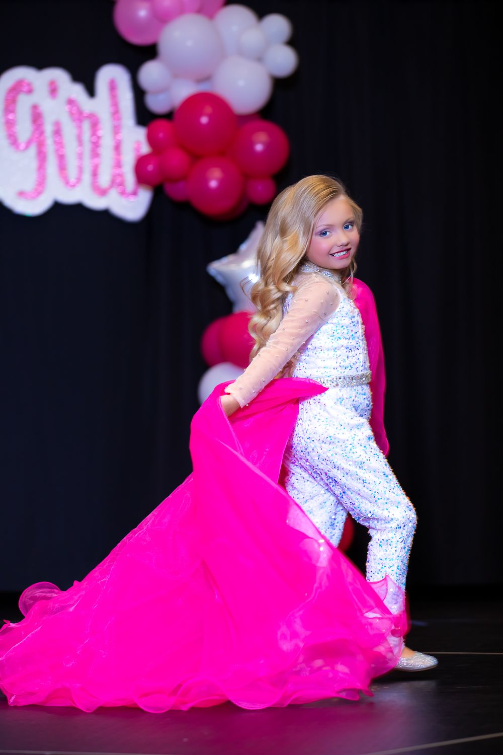 A performer in a white sequined outfit twirls with a flowing hot pink cape on stage.
