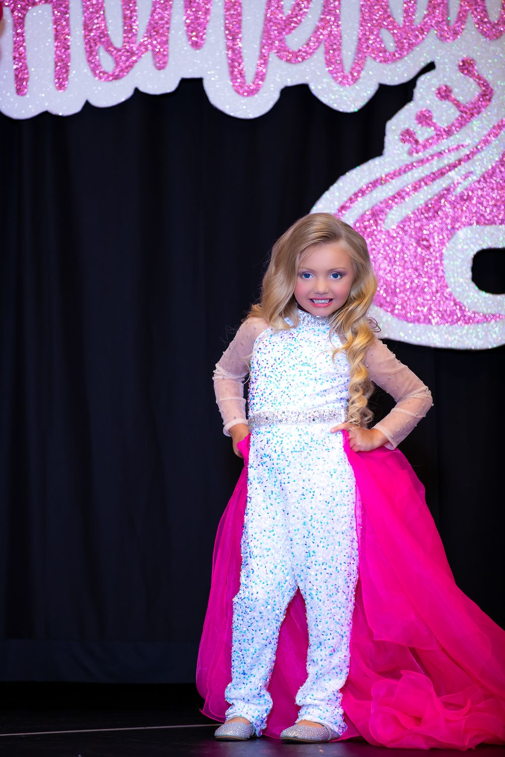 A performer poses confidently in a white sequined jumpsuit with pink cape on stage.