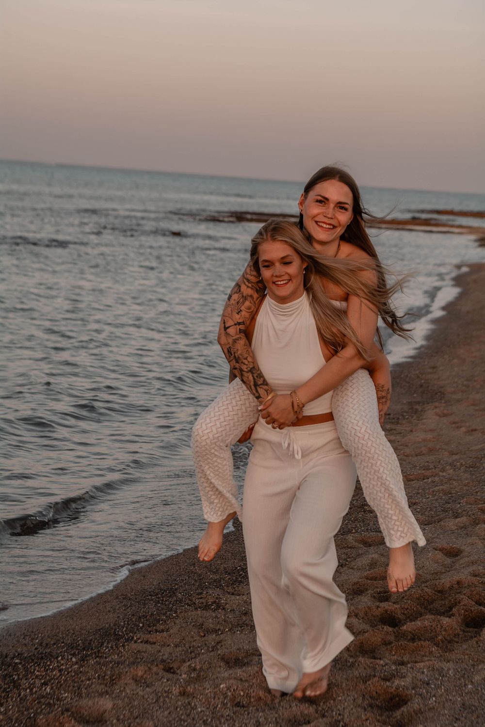 Two people in white outfits share a piggyback ride while walking along a beach at sunset.