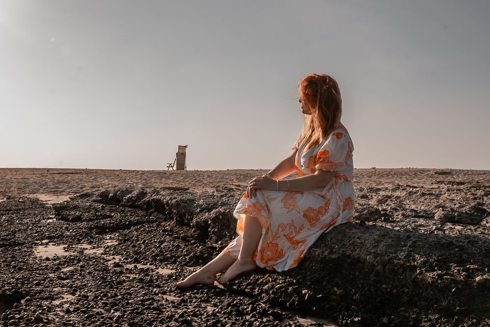 Figure in a flowy dress sits on rocky terrain at sunset while looking at distant silhouette on barren landscape.