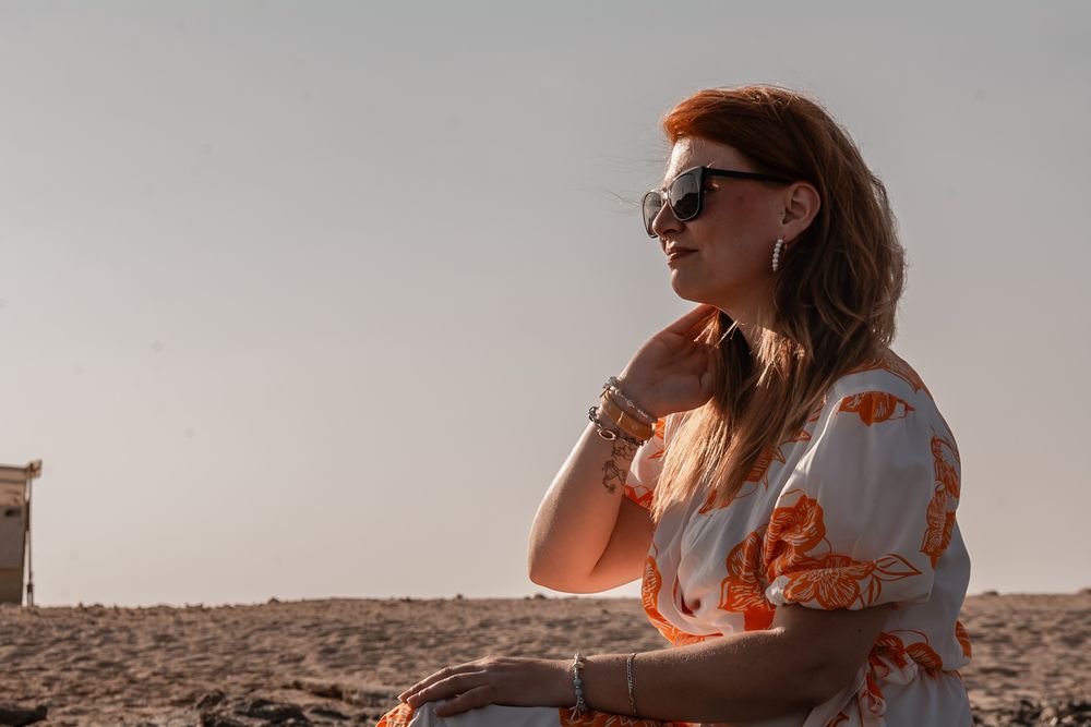 A silhouetted figure wearing sunglasses and floral dress sits thoughtfully on a sandy beach at sunset.