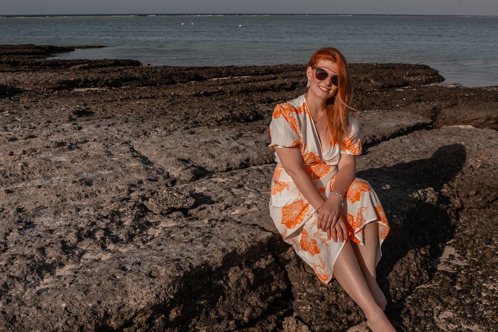 A person in an orange and white floral dress sits on rocky coastal terrain at sunset.