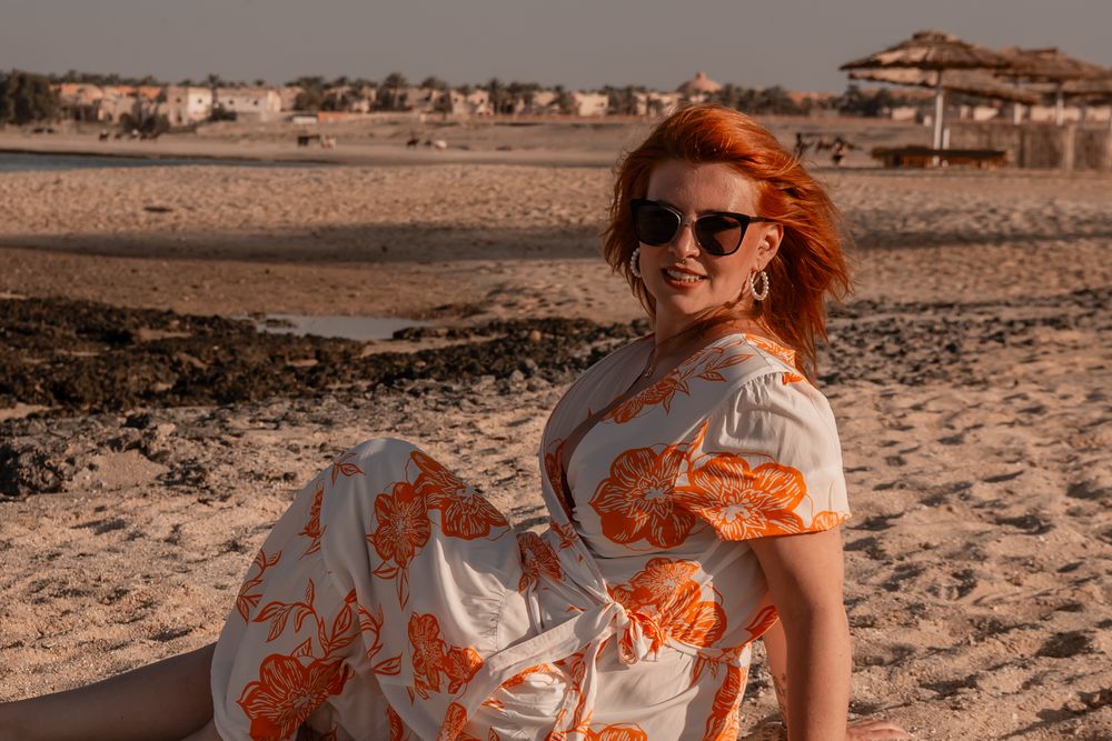 Person with red hair in flowy white dress poses in desert landscape at sunset while sand dances in the wind.