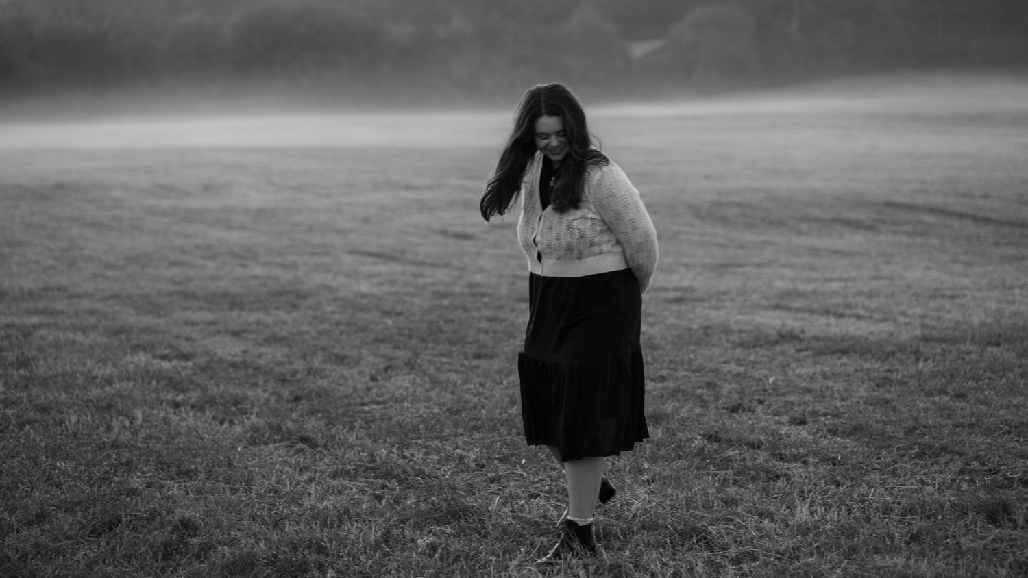 Ethereal black and white photo of a woman in a long skirt walking through a misty field at dusk.
