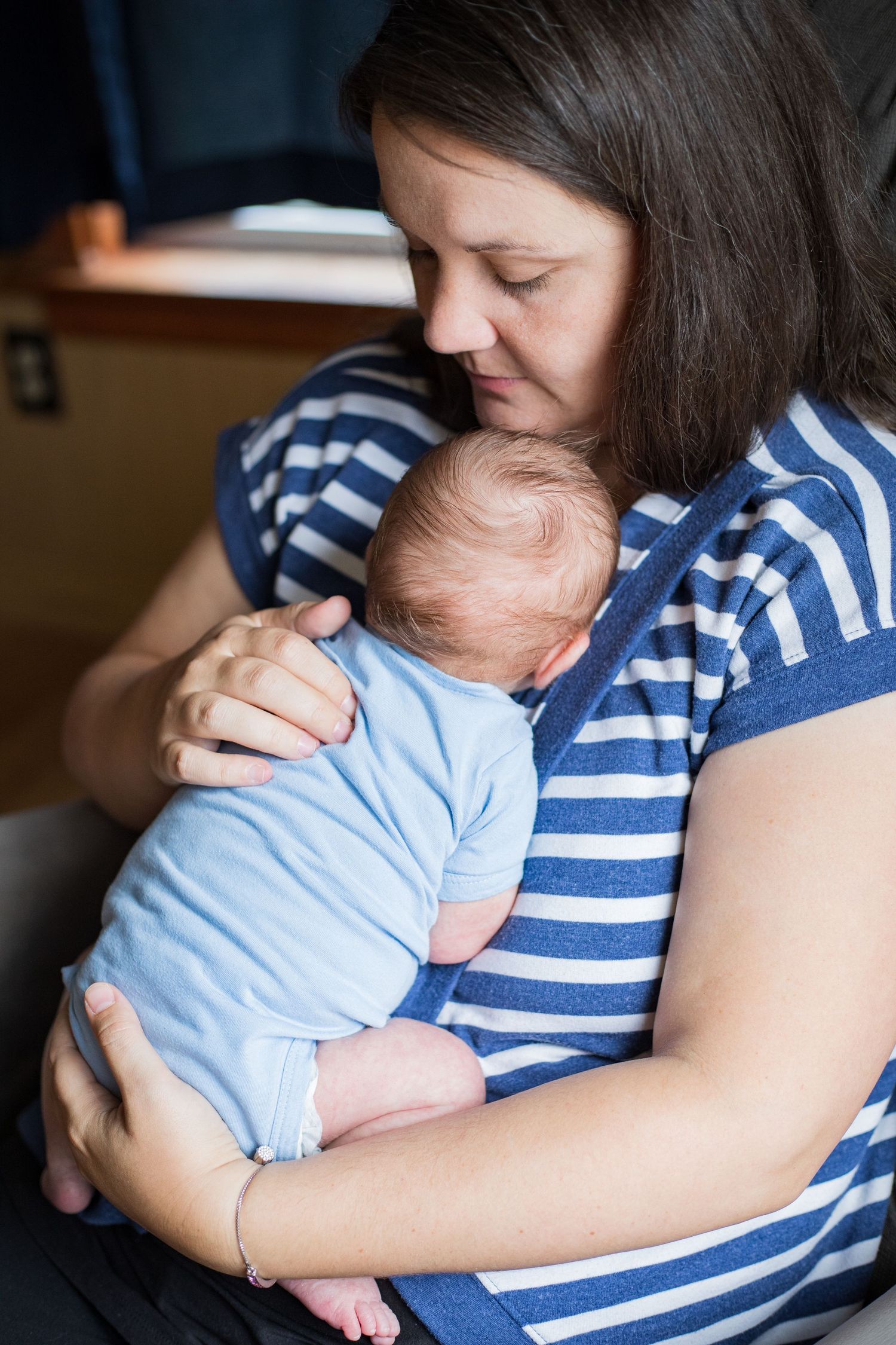 A dark-haired mother in a blue striped shirt tenderly holds her sleeping newborn against her chest.