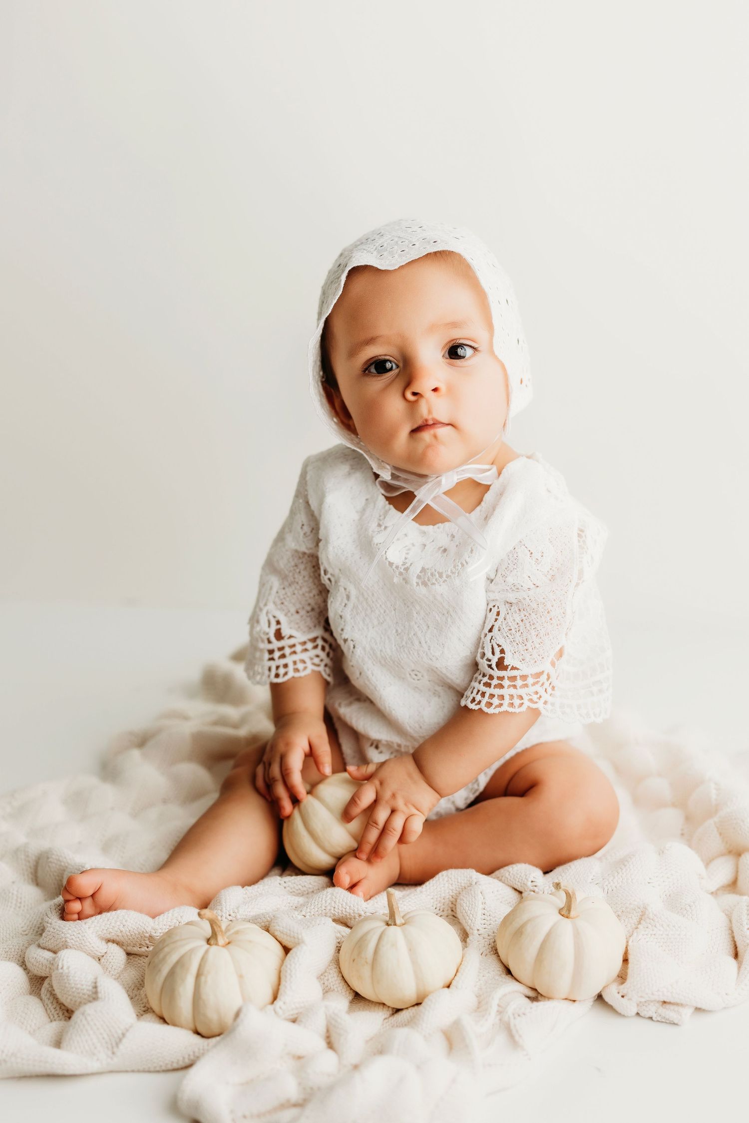 Baby in pink ruffled dress and bow sitting on white chair against brown background.