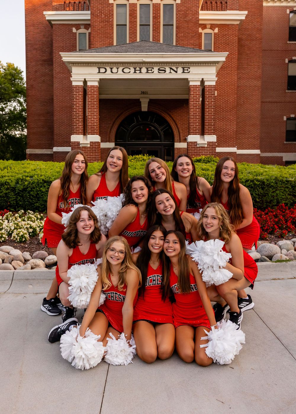 Group of cheerleaders in red uniforms with white pom poms pose outside brick Duchesne building entrance in Omaha, NE