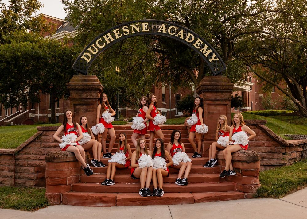 Dance Team in red and white uniforms sit on stone steps under an arched 'Duchesne Academy' entrance sign in Omaha, NE