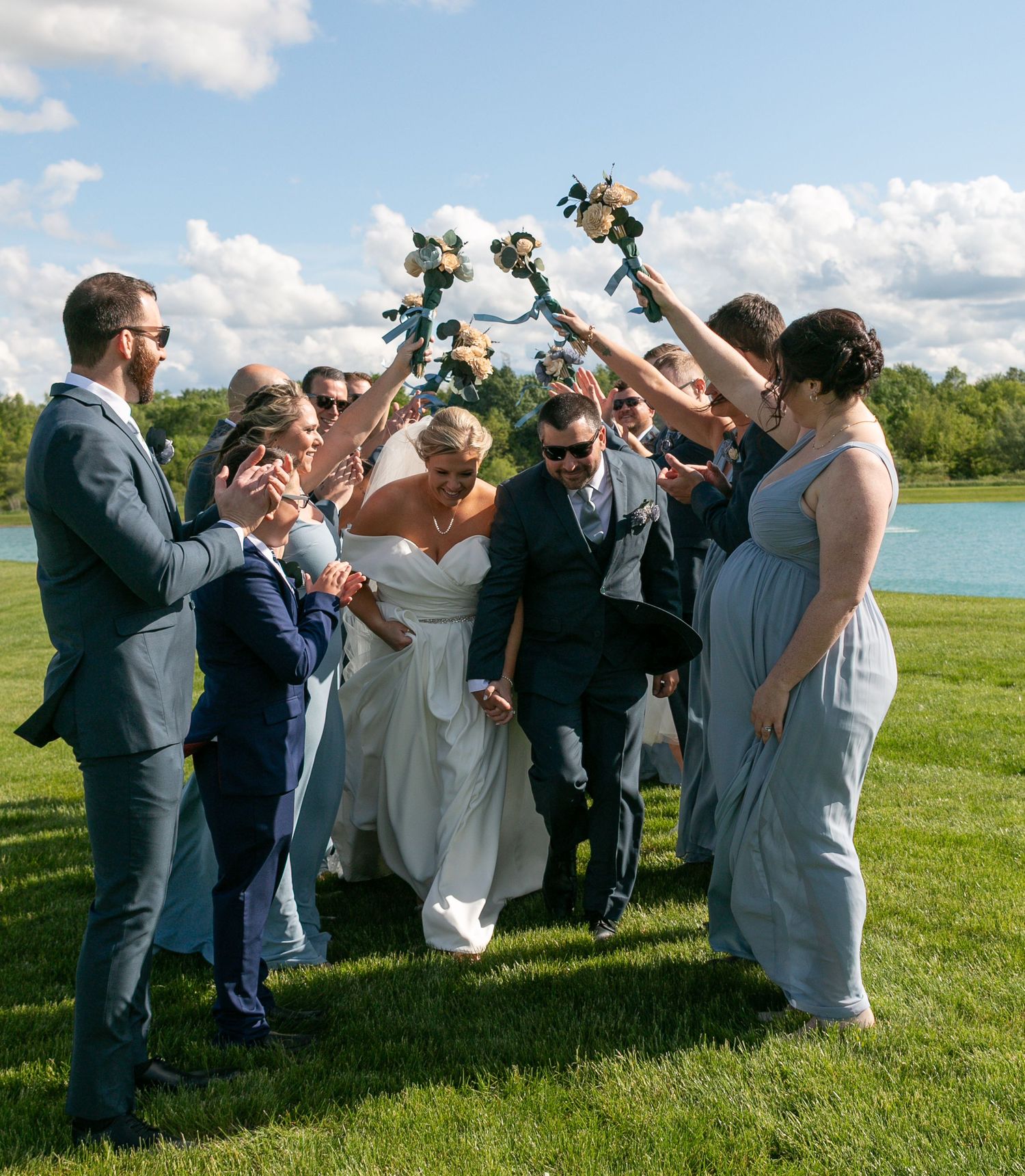 Wedding party celebrating on a grassy lawn next to a lake, raising bouquets in the air on a sunny day.