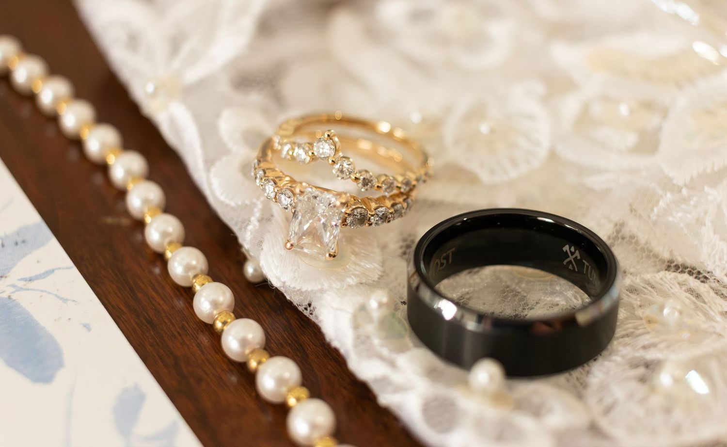 Gold diamond engagement ring and black wedding band displayed on lace with a string of pearls.