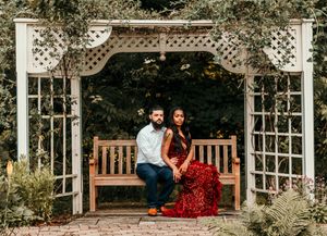 A couple sits on a wooden bench under an ornate white garden arbor with climbing vines in a romantic outdoor setting.