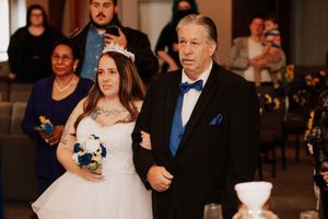 A traditional bride in a white wedding gown walks down the church aisle accompanied by a person in a blue tuxedo.