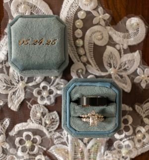 A vintage-style engagement ring with intricate detailing sits nestled in a pale blue velvet ring box against a floral lace background.