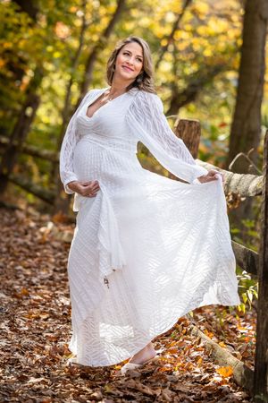 Pregnant woman in white lace dress posing in autumn forest, smiling and holding her belly among fallen leaves.