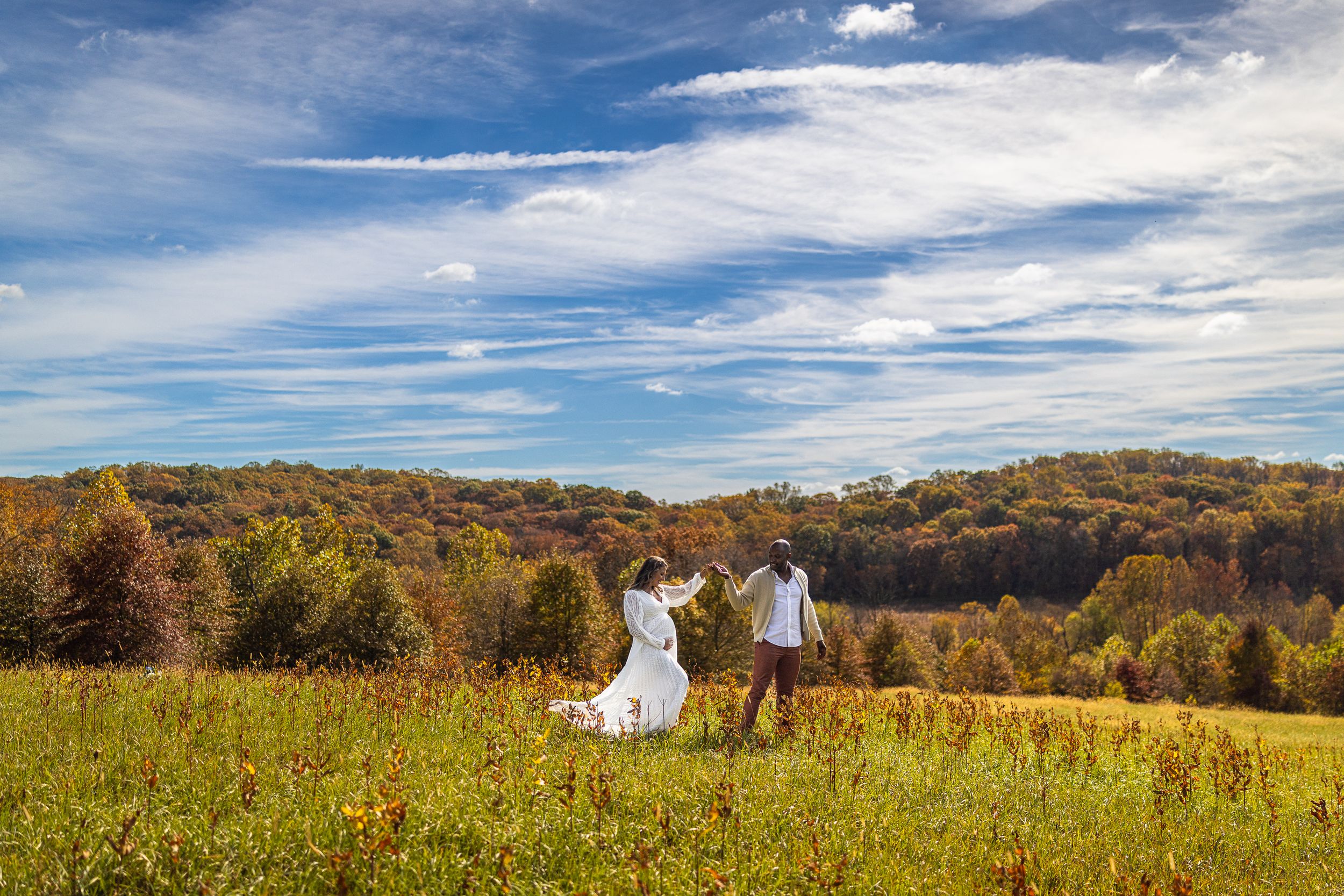 Bride and groom holding hands in a lush green meadow with colorful autumn trees and a dramatic blue sky.