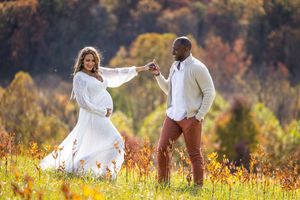 Pregnant couple posing in autumn field, woman in white flowing dress, man in white sweater holding hands outdoors.