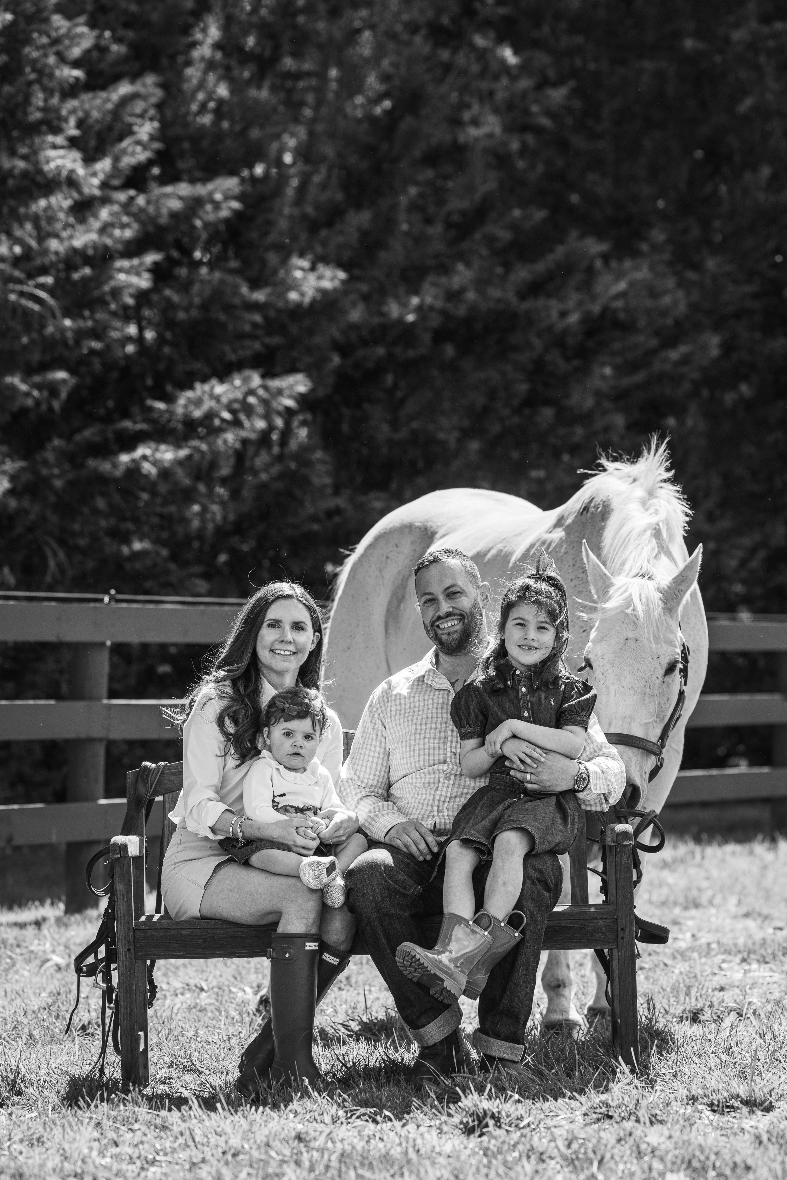 Black and white family portrait with parents, two children, and a white horse by a wooden fence outdoors.