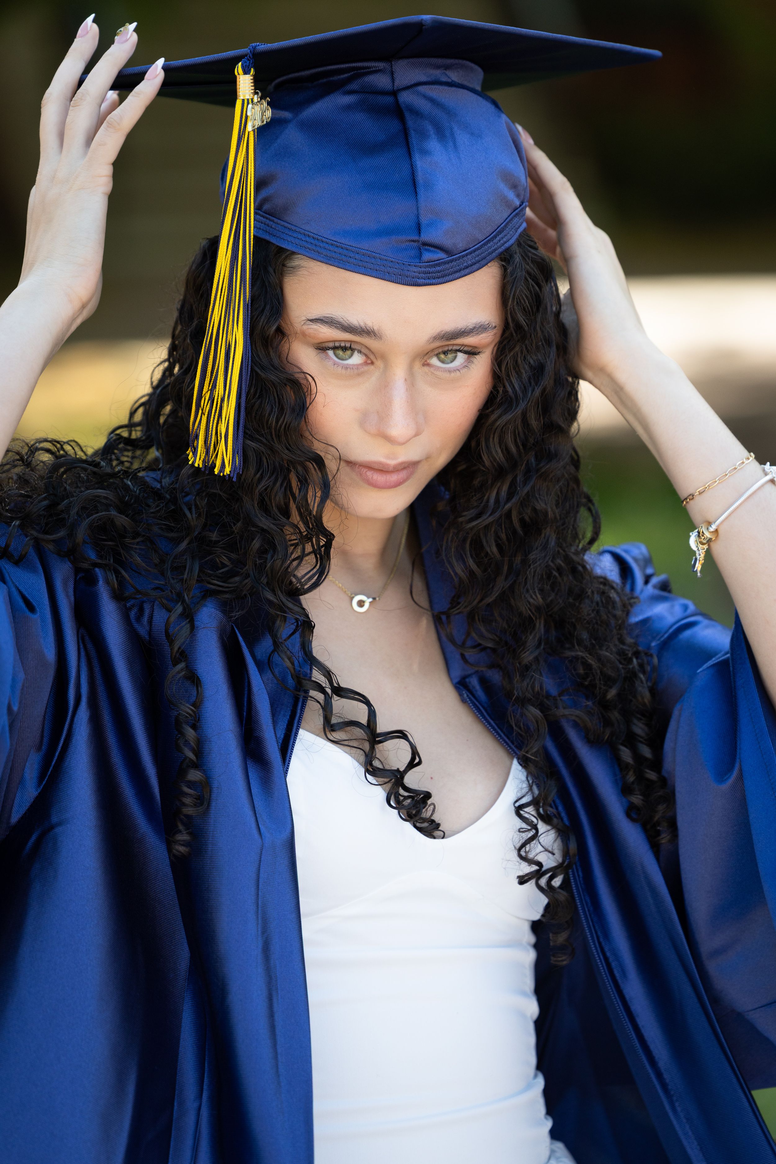 Young woman in blue graduation cap and gown adjusting her mortarboard with yellow tassel outdoors.