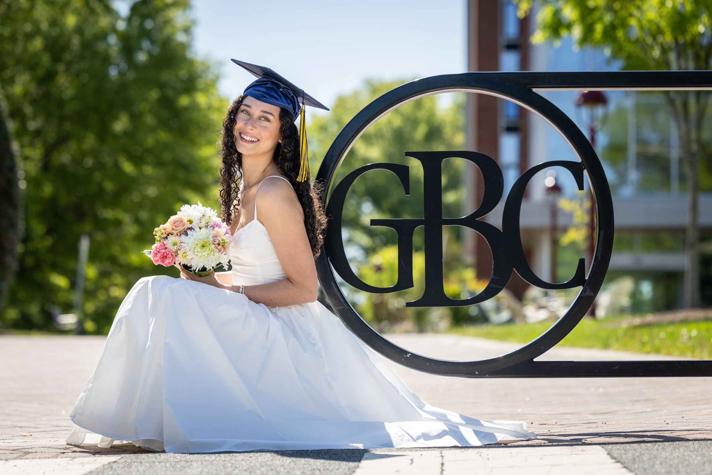 Smiling graduate in white gown and blue cap holds bouquet beside GBC sign outdoors on sunny day.