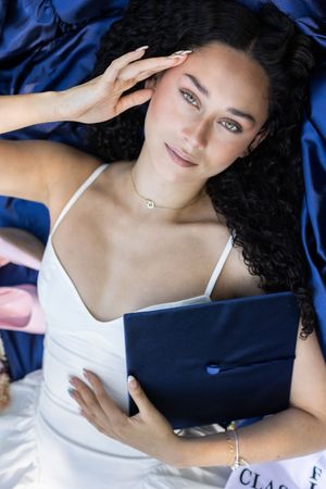 Young woman in white dress lying on blue satin, holding graduation cap, smiling at camera for senior portrait.