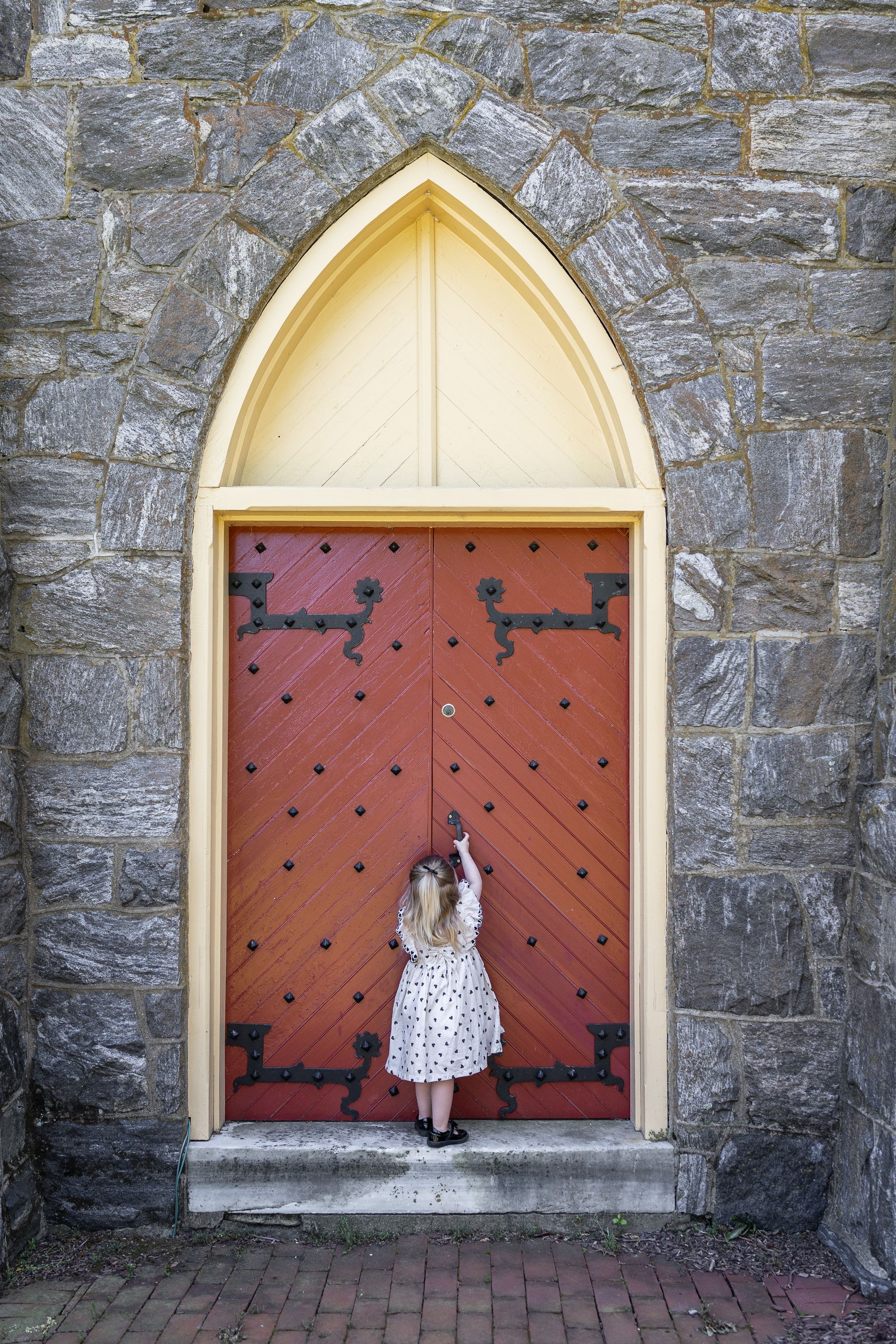 Young girl in floral dress knocking on large red Gothic arched door of stone church.