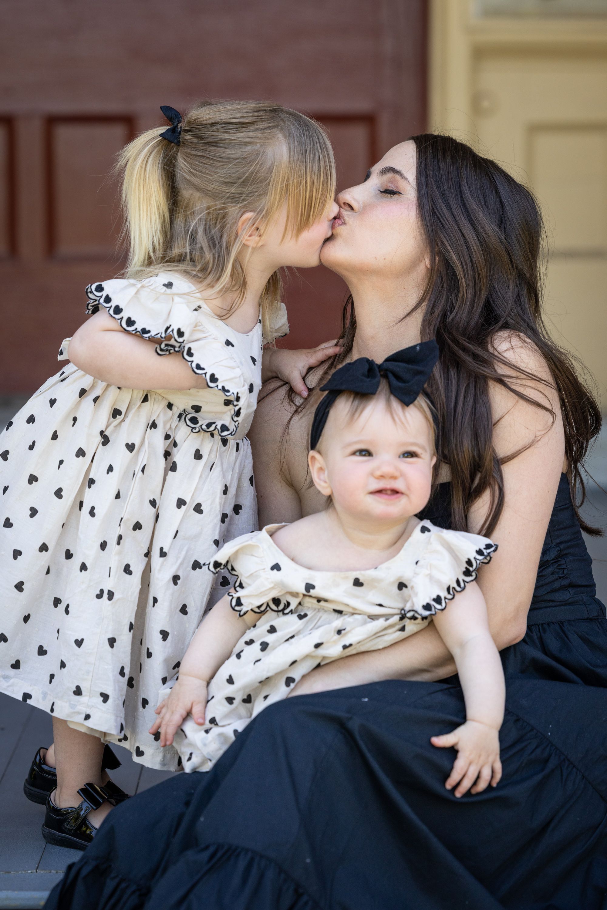 A mother in a black dress kisses her toddler daughter while holding her baby, both girls wearing matching heart-print dresses.