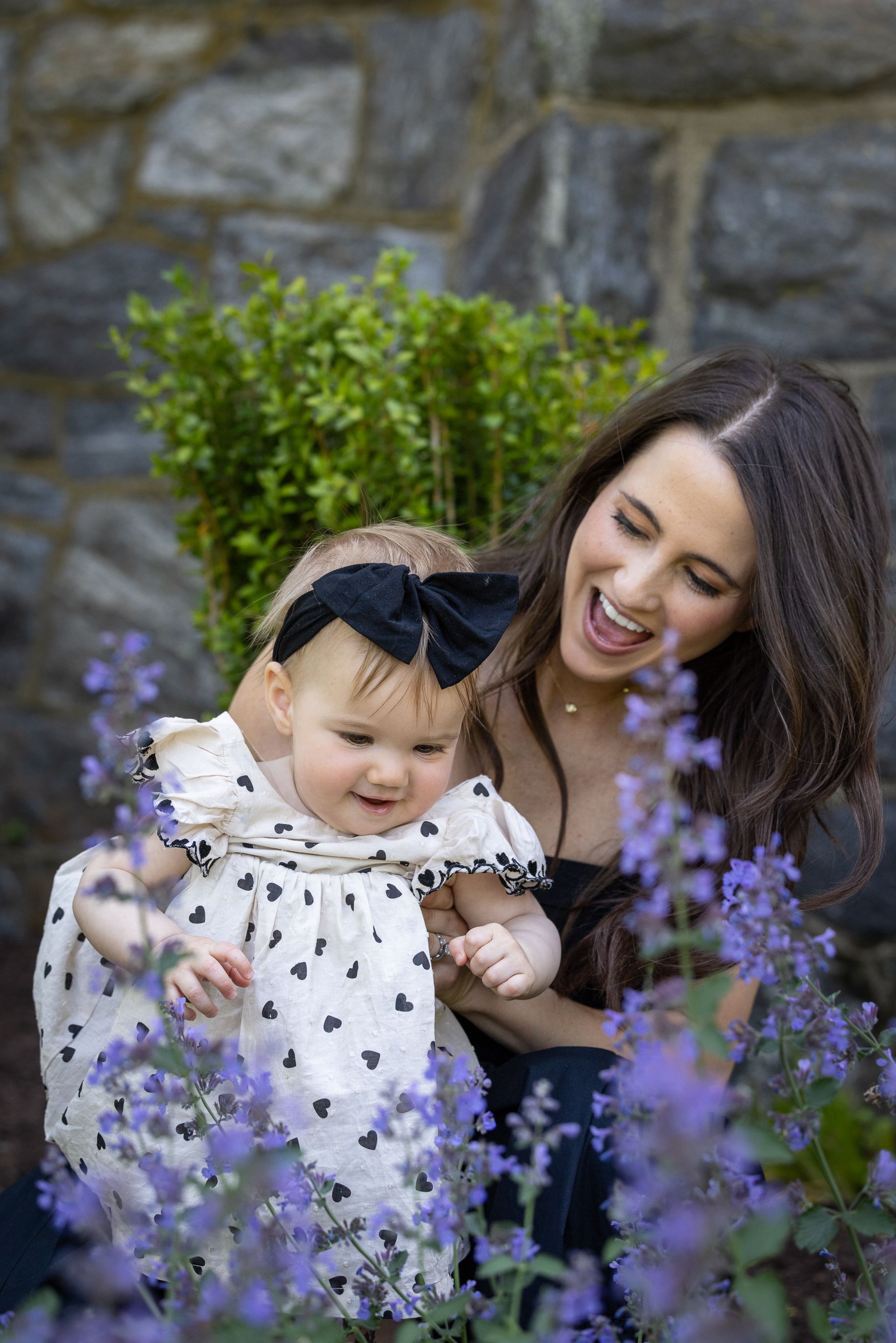 Smiling woman holding baby girl in heart-print dress with black bow headband among purple flowers.