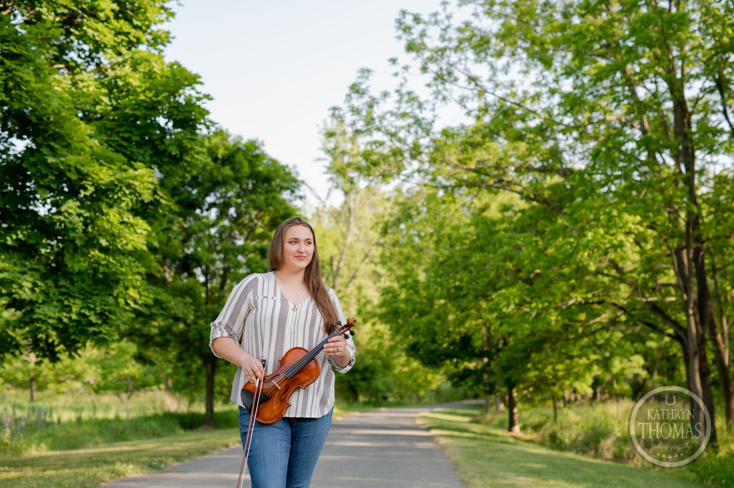 Musician stands on tree-lined path holding violin against natural green backdrop.