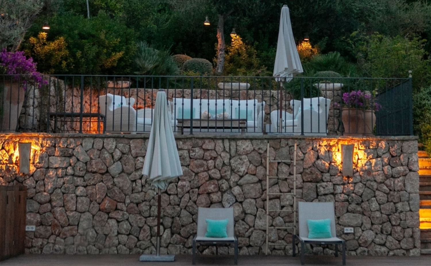 Stone wall terrace with illuminated lanterns and lounge chairs overlooking a pool area at night.
