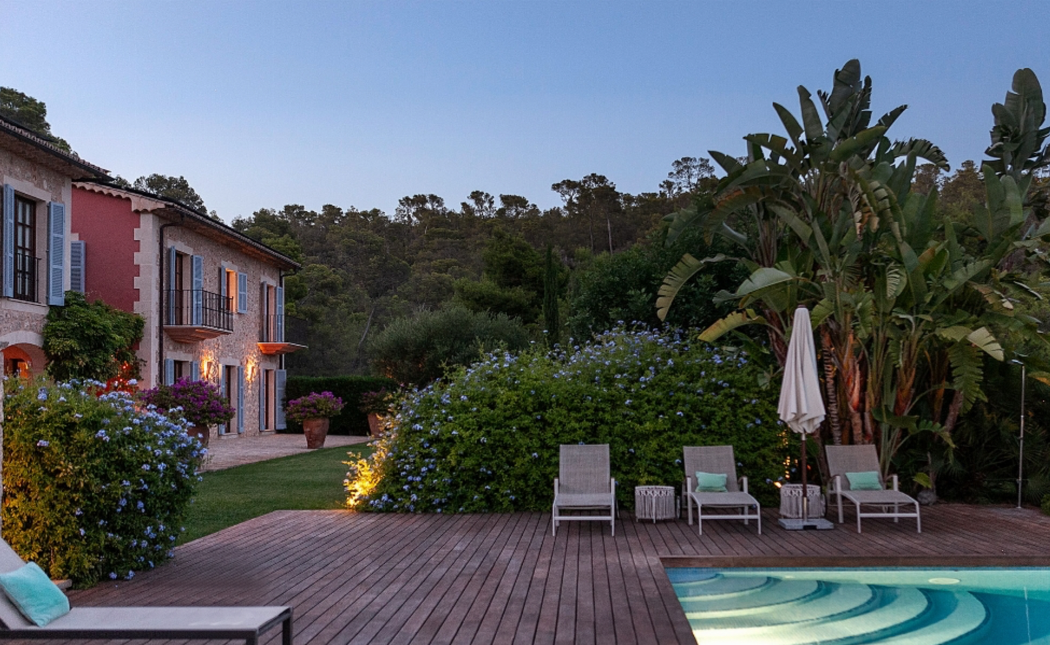 Luxury villa deck with loungers and swimming pool surrounded by palm trees and landscaping at dusk.