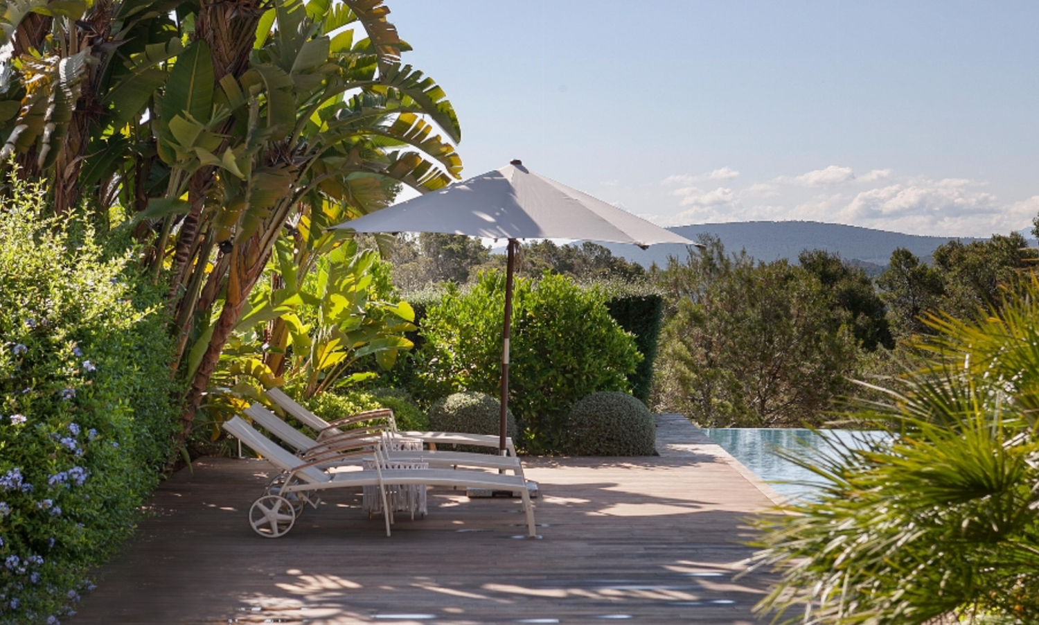 Pool loungers and umbrella on sunny terrace with mountain views and tropical landscaping.