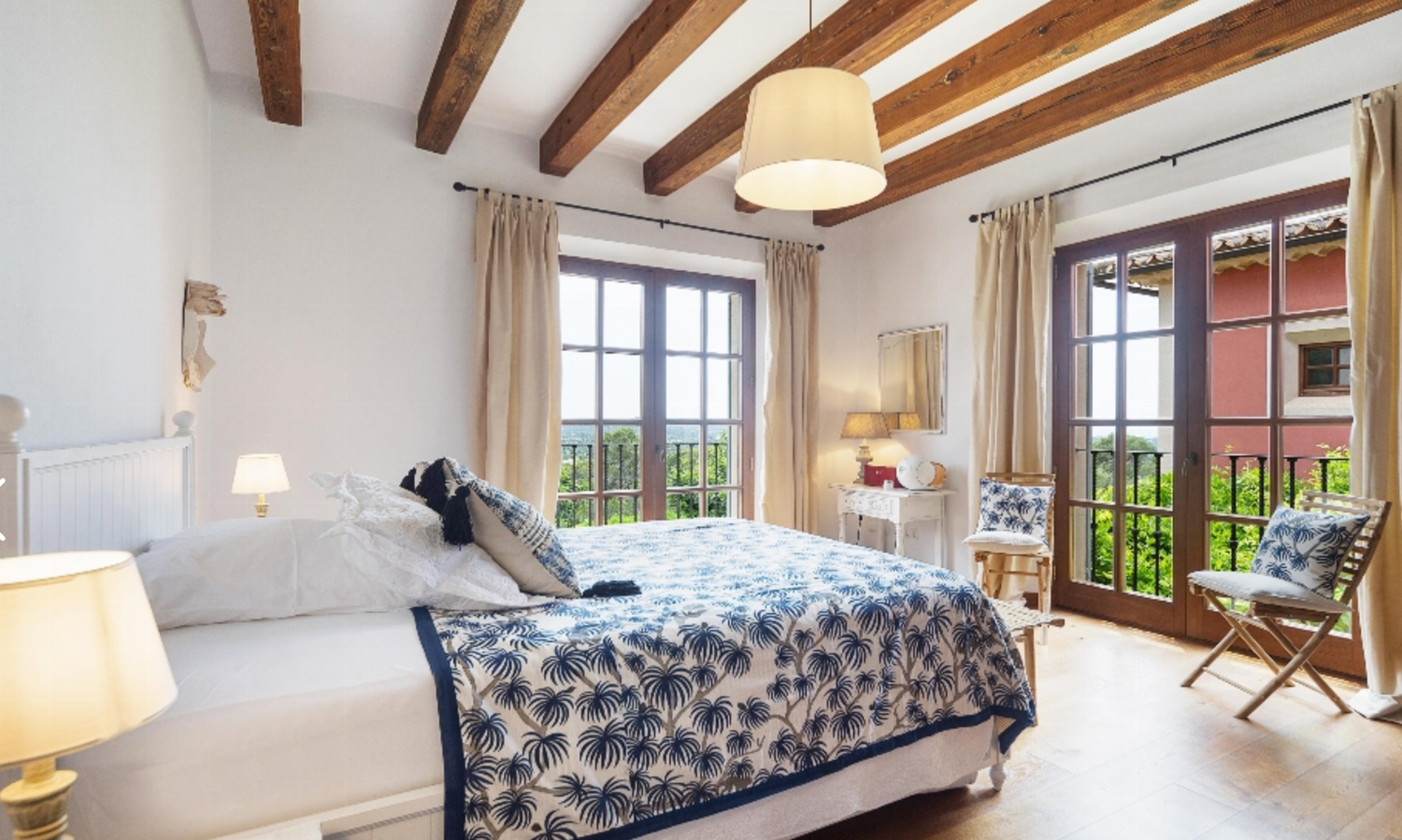 Bright bedroom with wooden ceiling beams, floral bedding, and balcony access through French doors.