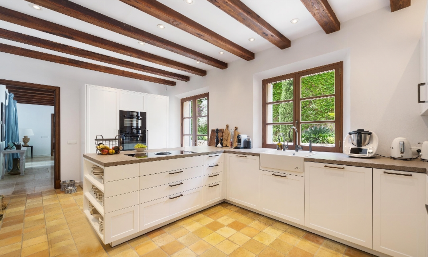 Modern white kitchen with exposed wooden ceiling beams, terra cotta floor tiles and wooden-framed windows.