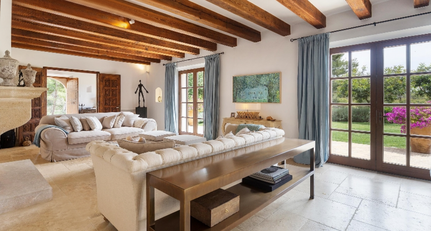 Spacious living area with exposed ceiling beams, white furniture, and glass doors leading to outdoor space.