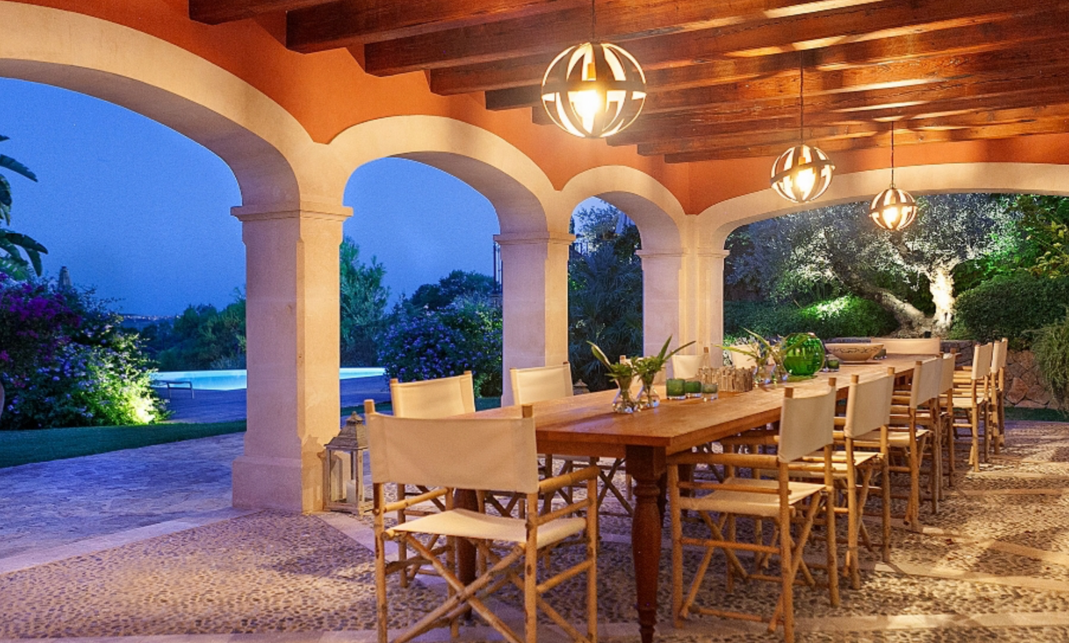 Covered outdoor dining area with wooden table and chairs under arched colonnade at dusk.