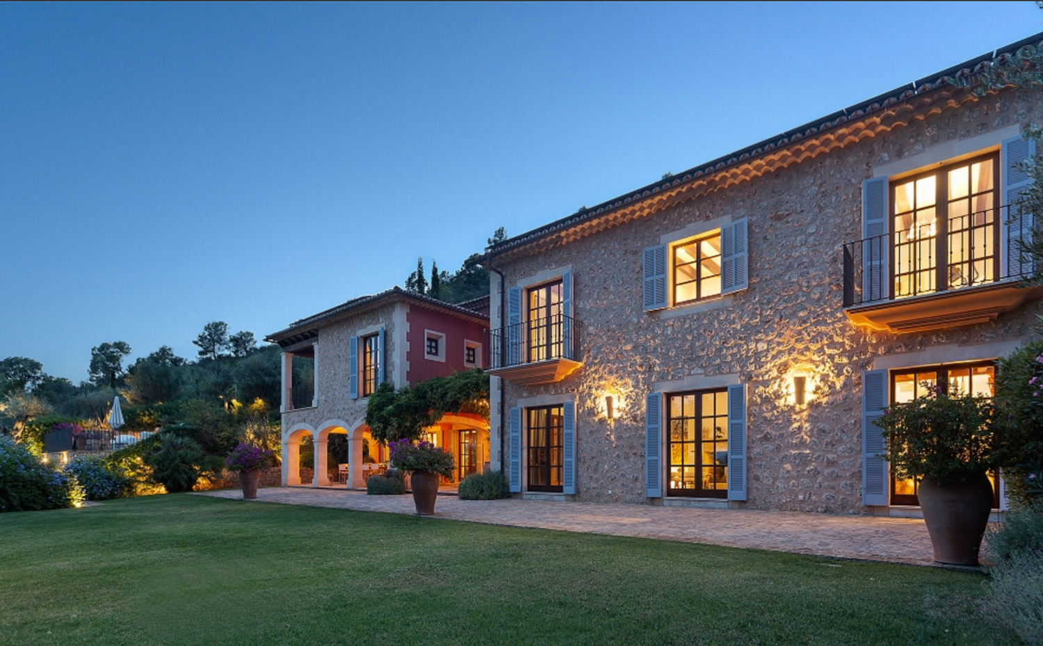 Stone villa with blue shutters and warm lighting glowing from windows at dusk.