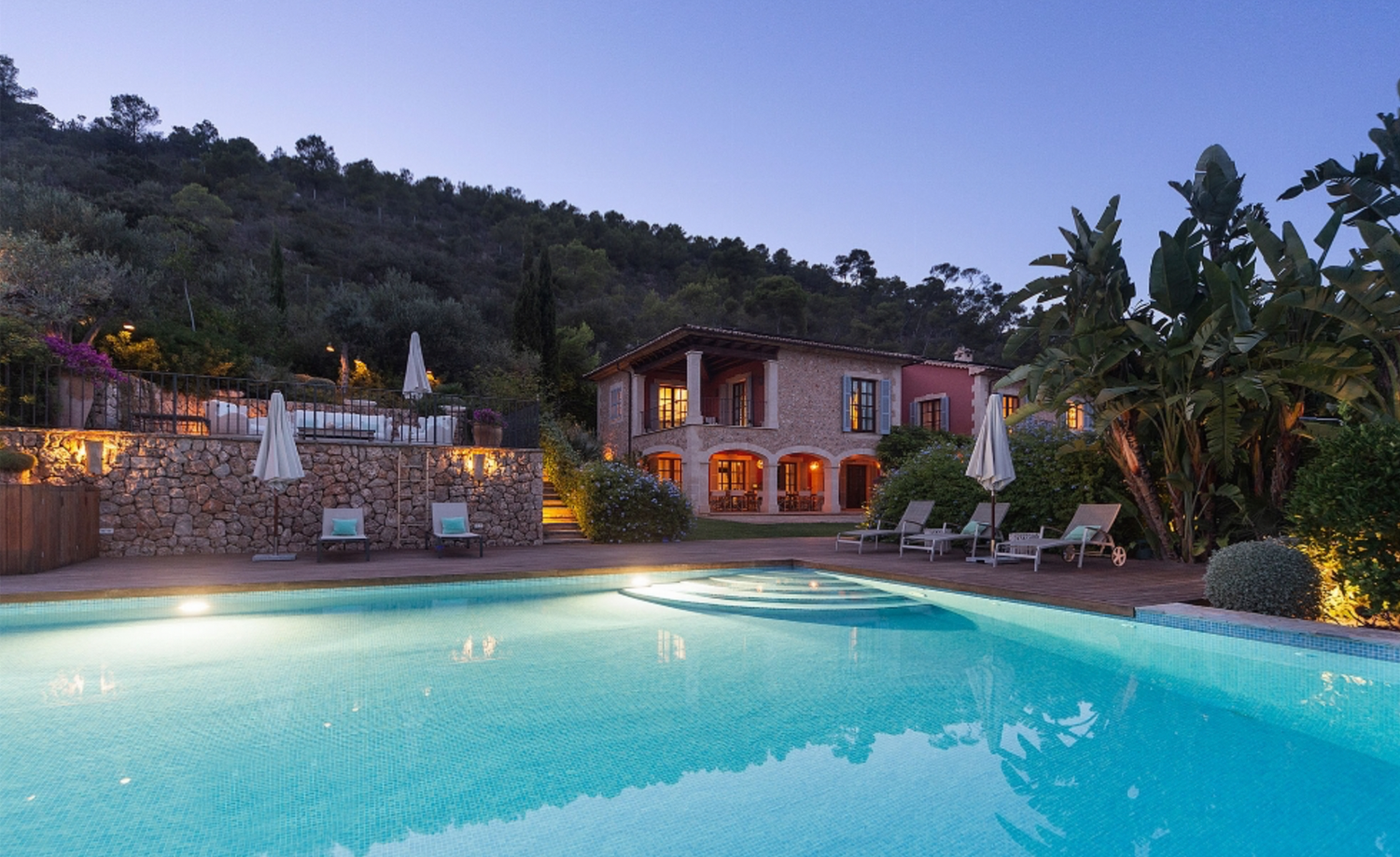 Twilight view of an illuminated villa with swimming pool and palm trees.