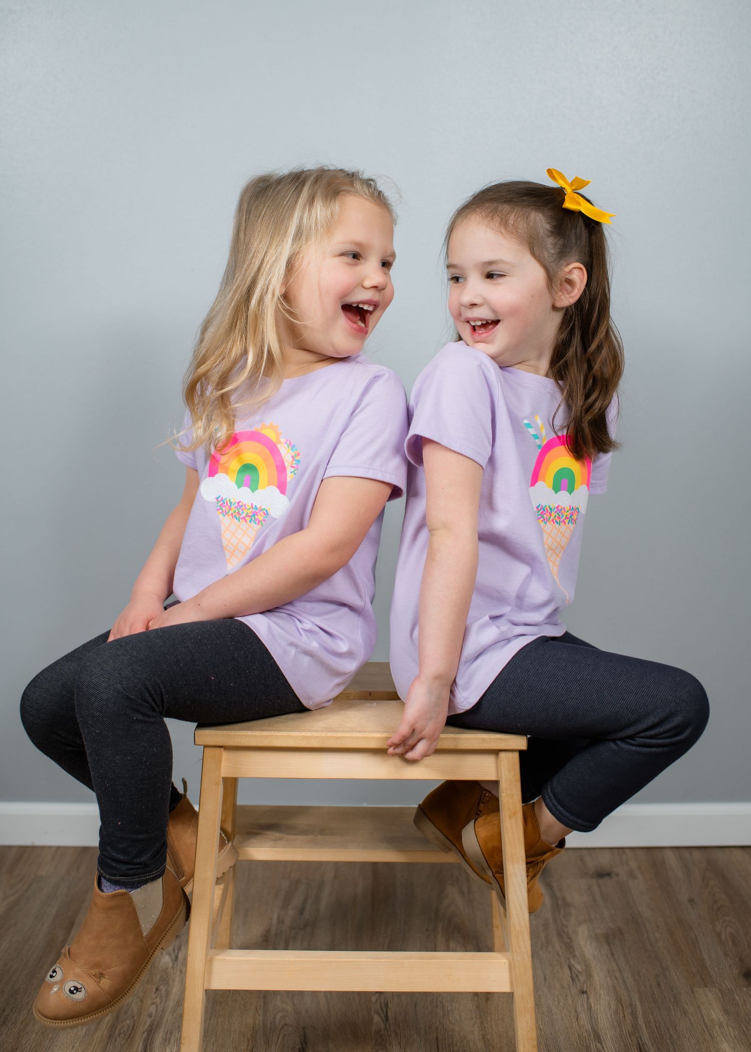Two children in matching purple rainbow shirts laugh while sitting on a wooden stool.