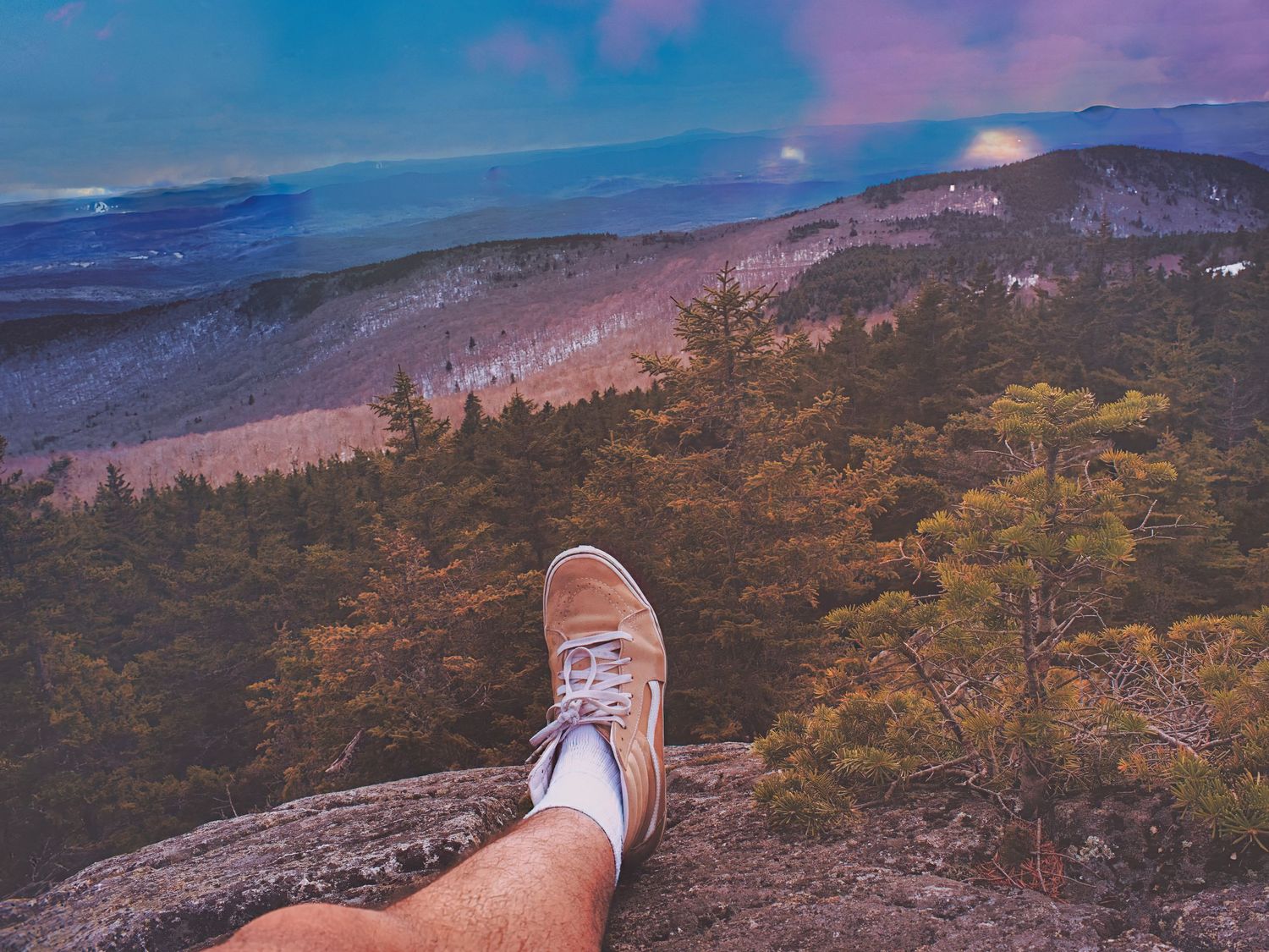 A hiker rests their sneaker-clad foot on a rocky mountain overlook with a scenic forested valley below.