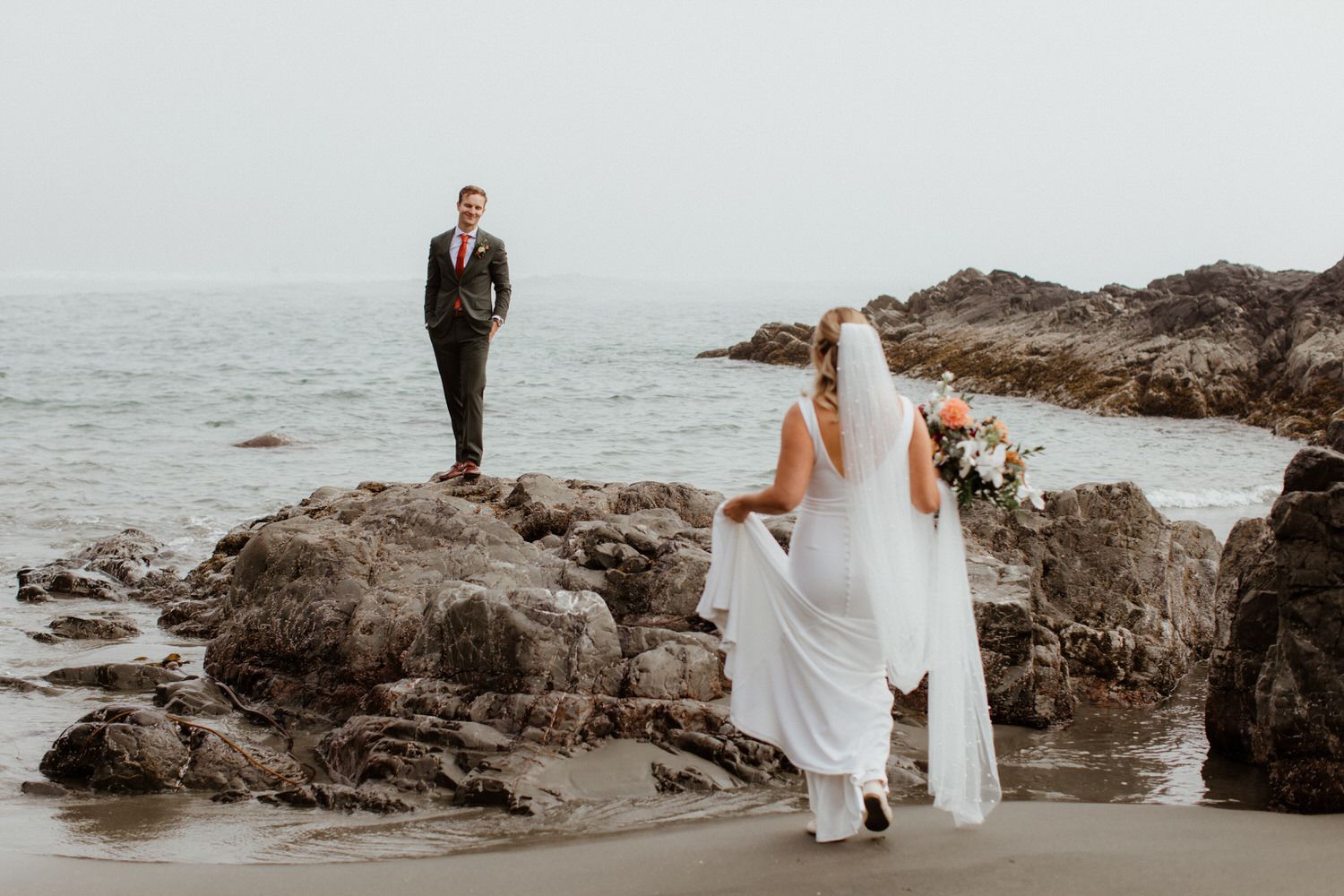 Bride in white gown and veil walks toward groom standing on coastal rocks by the ocean.