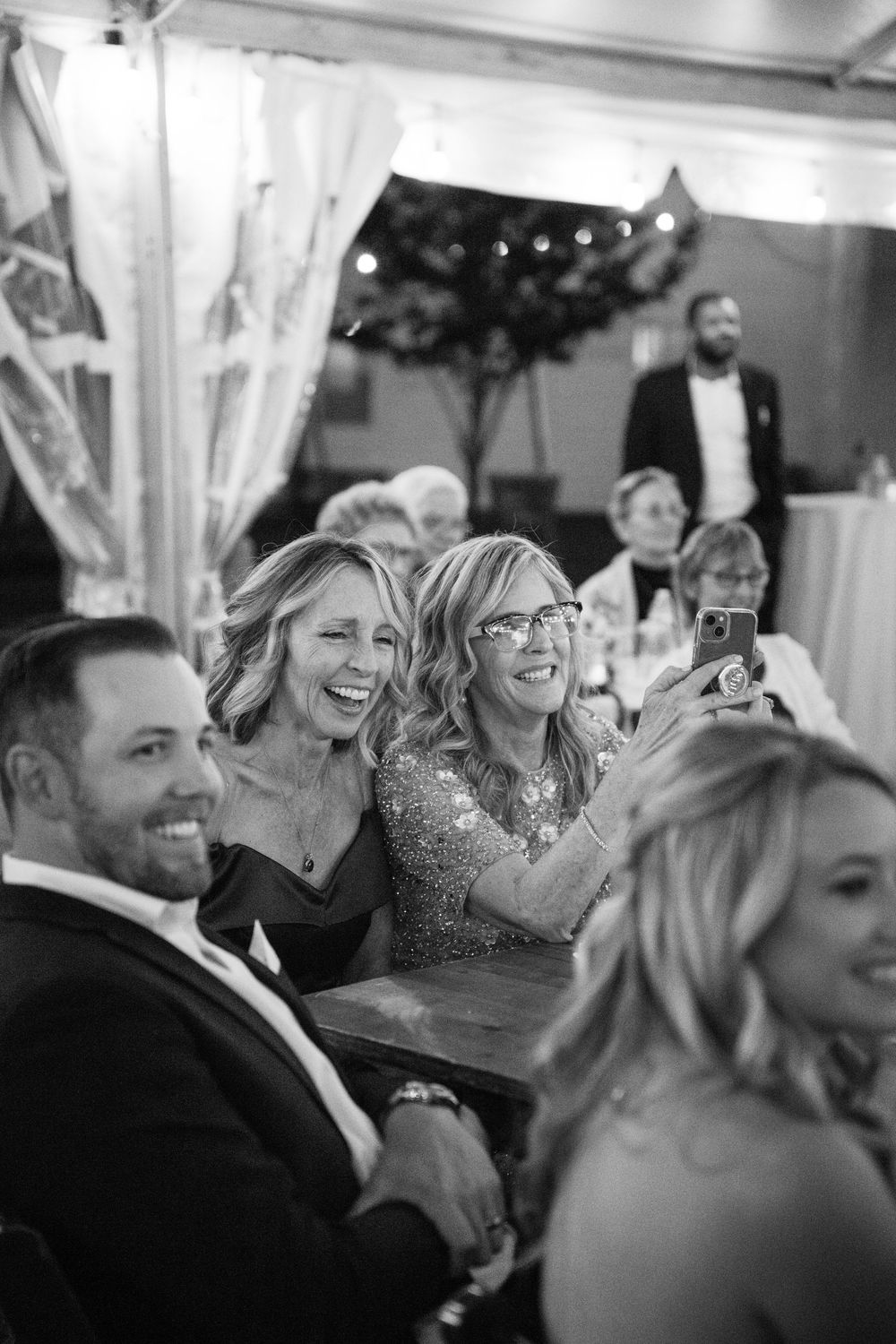 Black and white photo of guests laughing and having fun during a wedding reception with decorative lights in background.