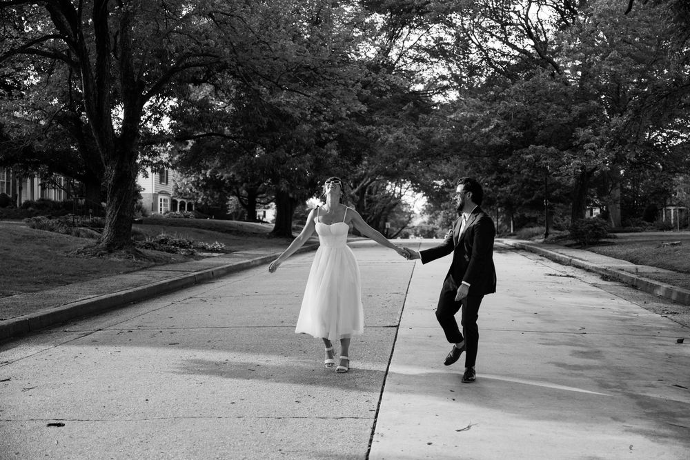A couple in formal attire dances together on a tree-lined street in black and white photography.