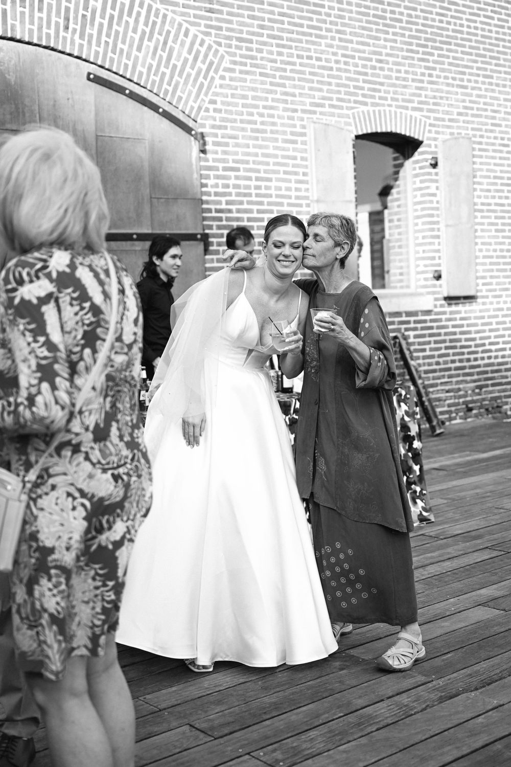 A candid black and white photo of wedding guests gathered on a wooden deck against a brick wall.