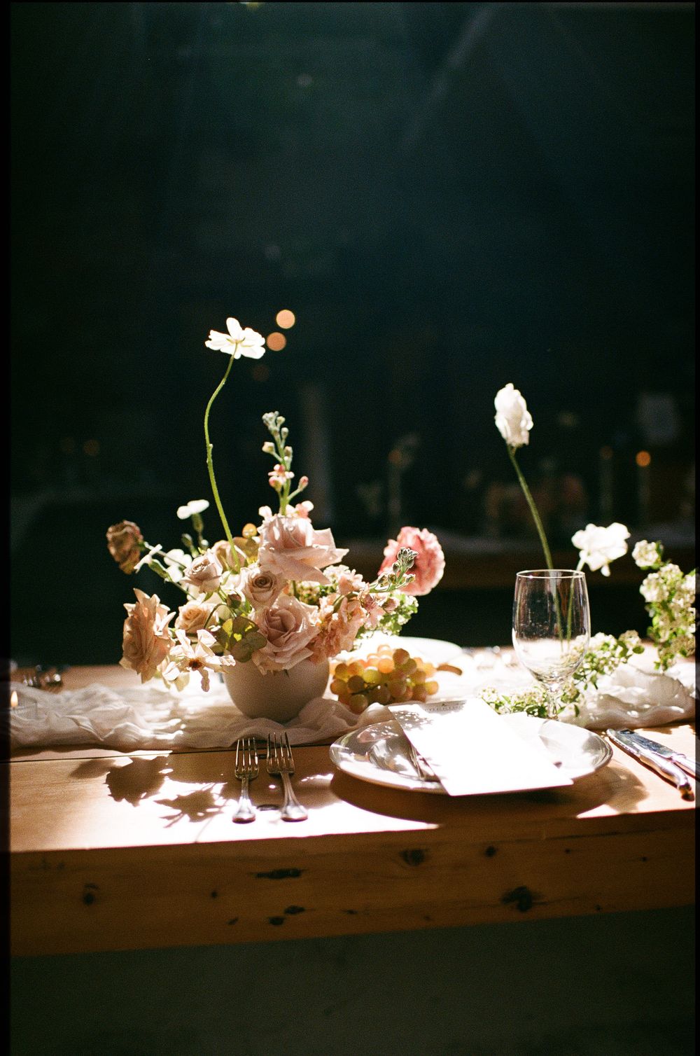 An artful arrangement of delicate flowers and decorative elements displayed on a rustic wooden table lit by soft spotlighting.