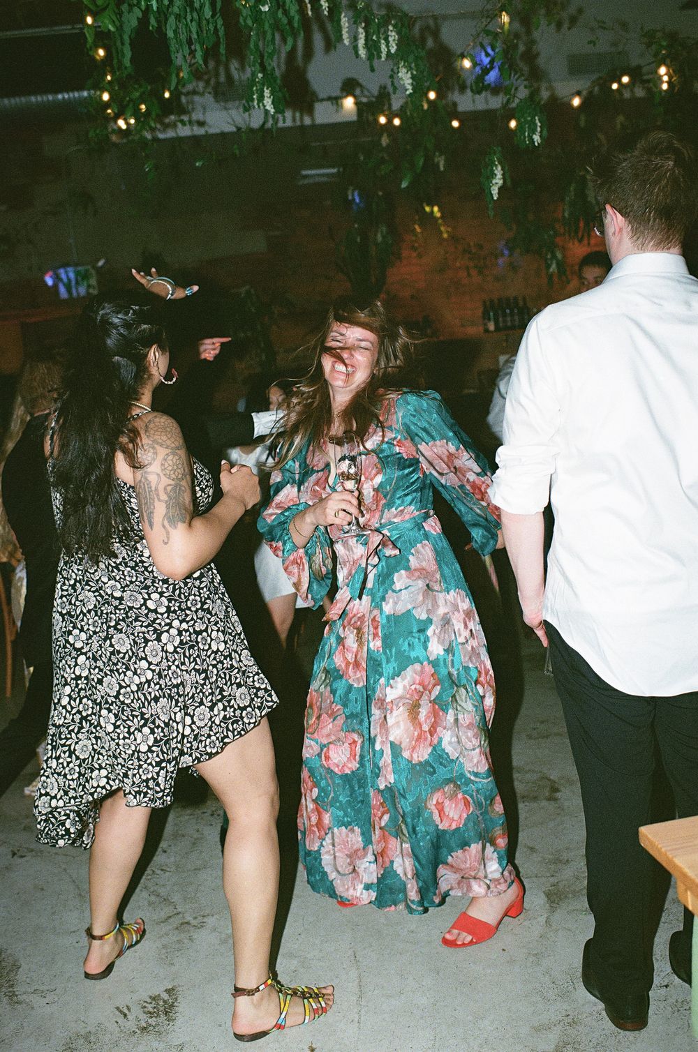 Two people dance together at a party, one in a floral maxi dress and one in a polka dot dress with party lights in background.