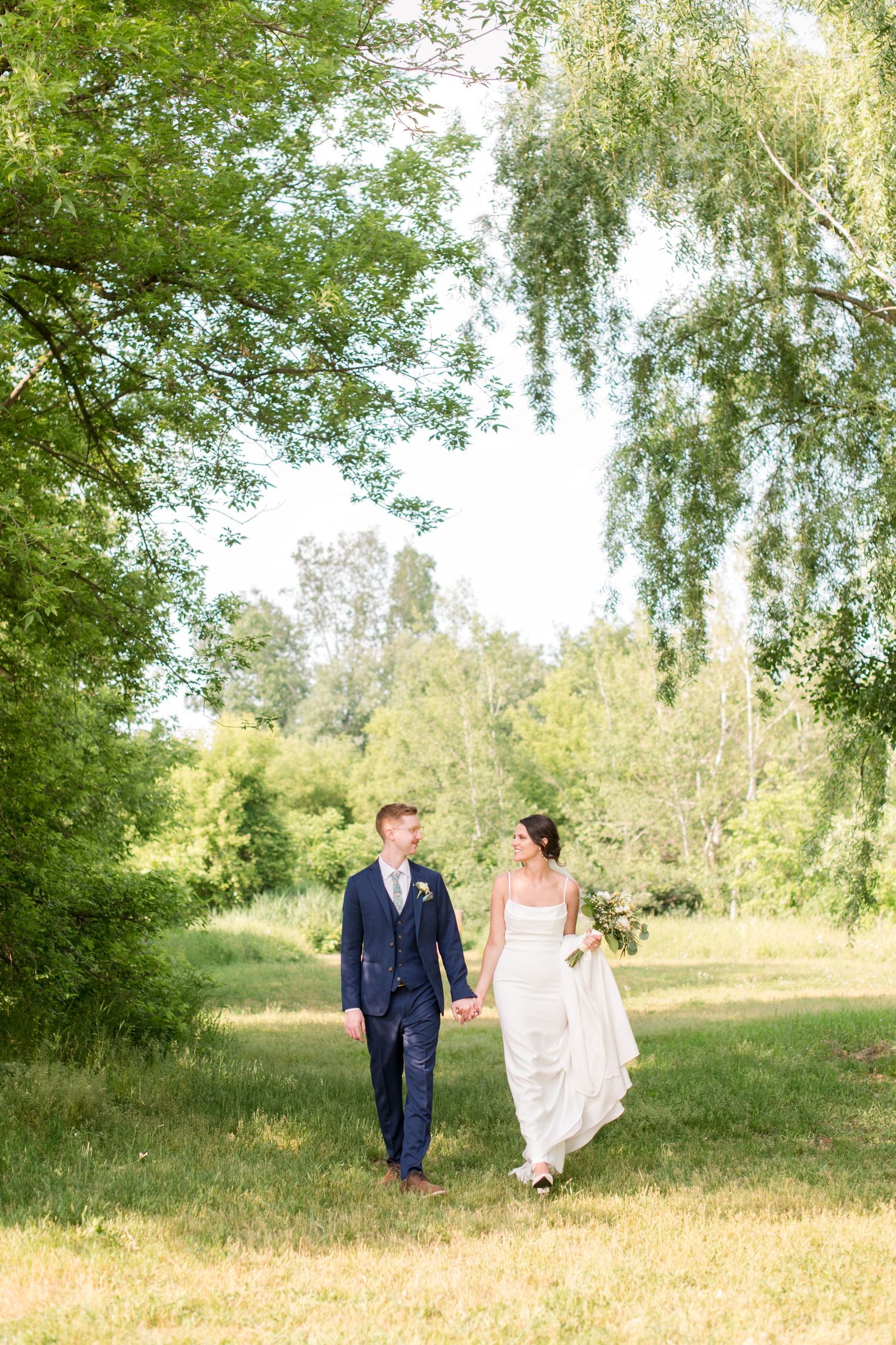 Bride and groom walk hand in hand through lush green park during summer wedding photo session.